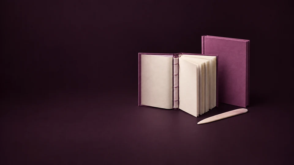 An open orchid casebound book exposes cream pages and a sewn spine beside a separate hardcover shell and bone folder. - Printing & Publishing Services Meta ads research index
